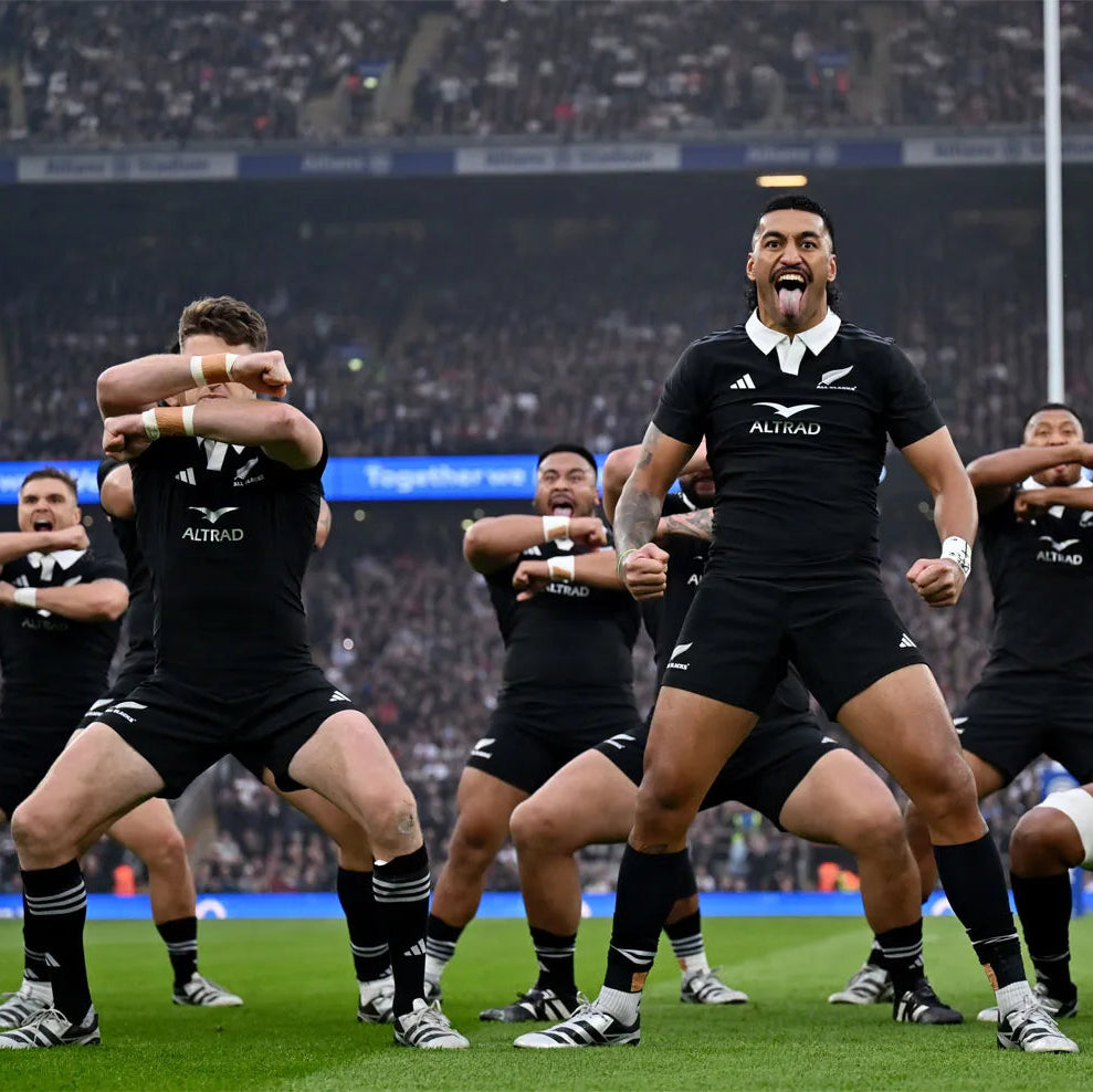 New Zealand rugby players performing the haka on a sports field with a crowd in the background