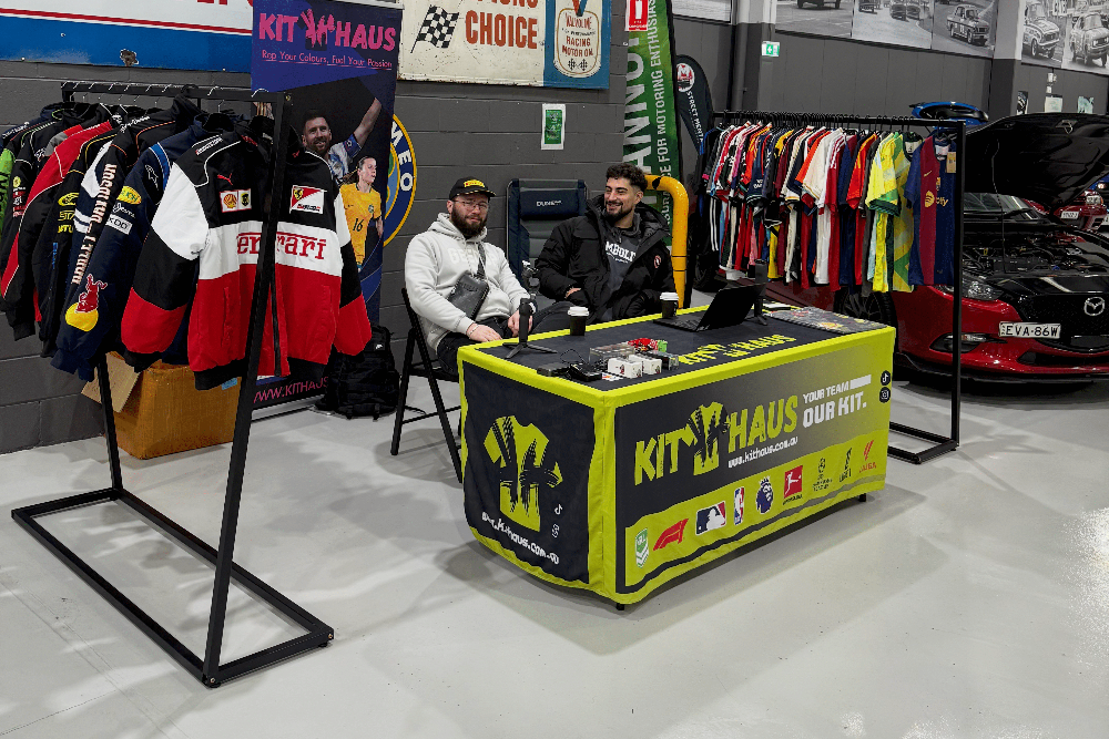 Two men sitting at a table with racing gear and branded clothing in a warehouse setting.